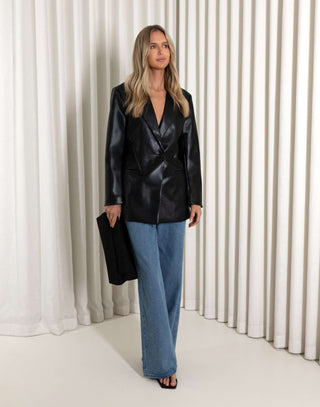 Woman wearing a black leather jacket and blue jeans standing against a white curtain.