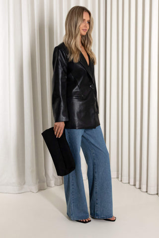 A woman models a black faux-leather blazer with a V-neckline, paired with wide-leg blue jeans and holding a black clutch, set against a background of white vertical drapes.