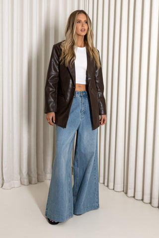 A woman modeling a dark brown faux-leather blazer over a white crop top and light blue wide-leg denim jeans, standing in front of a white pleated curtain backdrop.