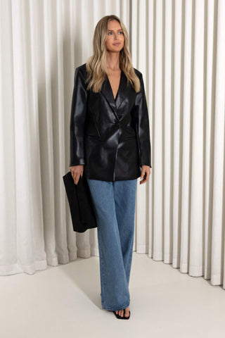 A woman stands in front of white pleated curtains wearing a black leather-look blazer, wide-leg blue jeans, and black sandals, while holding a black clutch.