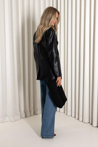 A woman stands in profile wearing a black leather jacket, blue wide-leg jeans, and black heels, set against a background of white vertical drapes.