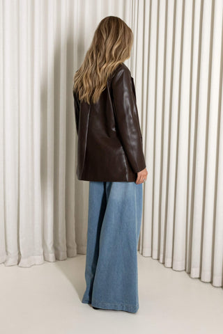 A woman stands with her back to the camera wearing an oversized dark brown leather jacket and wide-leg blue jeans, posed in front of a cream-colored pleated curtain backdrop.