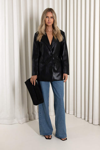 A woman stands in front of a white draped curtain backdrop wearing an oversized black faux-leather blazer, blue wide-leg jeans, and black sandals, while holding a black clutch.