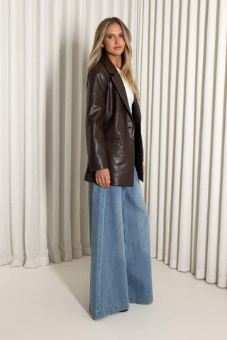 A woman modeling a dark brown faux-leather blazer over a white top and wide-leg blue jeans, standing in front of a neutral cream-colored pleated curtain backdrop.