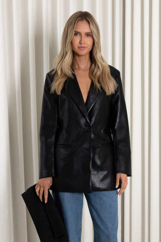 A woman with blonde hair wearing a black faux-leather blazer and blue jeans, holding a matching black blazer against a neutral cream-colored curtain backdrop.