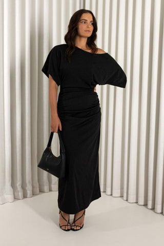 A woman modeling a black off-the-shoulder maxi dress with a ruched waist, holding a black handbag against a white pleated curtain backdrop.