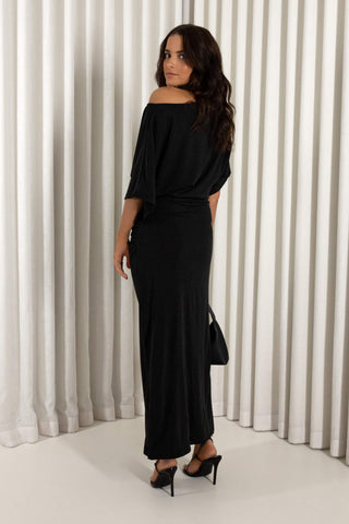 A woman modeling a black off-the-shoulder maxi dress with elbow-length sleeves, shown from the back against a white pleated curtain backdrop.