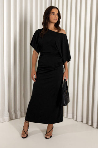 A woman modeling a black off-the-shoulder midi dress with a gathered ruched waist, paired with black strappy heels and holding a black handbag, set against a white pleated curtain backdrop.