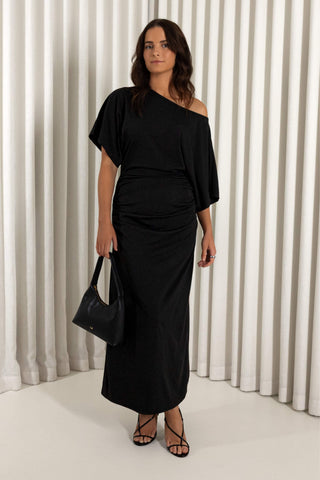 A woman modeling a black one-shoulder midi dress with draped short sleeves and a gathered waist, paired with black strappy heels and a black handbag, set against a white pleated curtain backdrop.