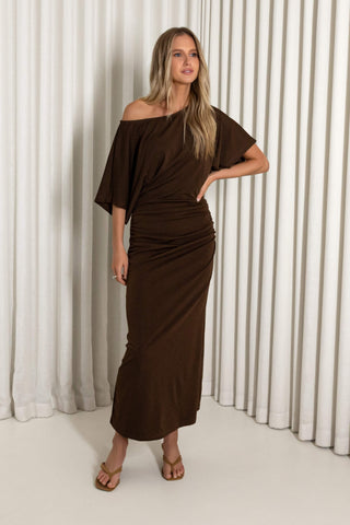 A woman modeling a dark brown off-the-shoulder midi dress with draped, gathered detailing, standing against a white curtain backdrop.
