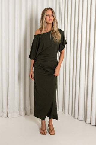 A woman modeling an olive green off-the-shoulder midi dress with draped short sleeves and a gathered waist, standing against a white curtain backdrop.