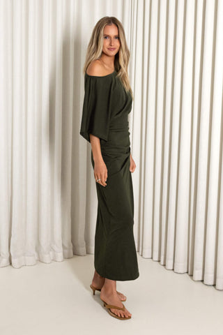 A woman modeling an off-the-shoulder dark olive green midi dress with draped sleeves and a gathered skirt, standing against a background of white vertical drapes.