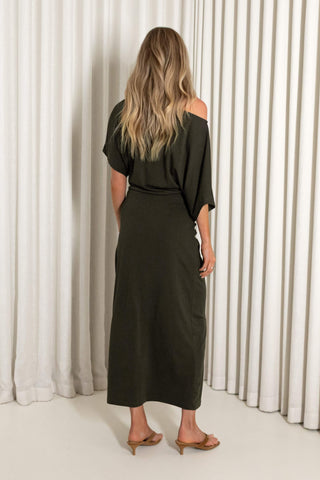 A woman viewed from behind wearing an olive green off-the-shoulder midi dress paired with tan heeled sandals, standing in front of white pleated curtains.