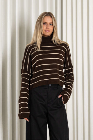 A woman wearing a cropped brown knit turtleneck sweater with horizontal cream stripes and black wide-leg pants, standing against a white pleated curtain backdrop.