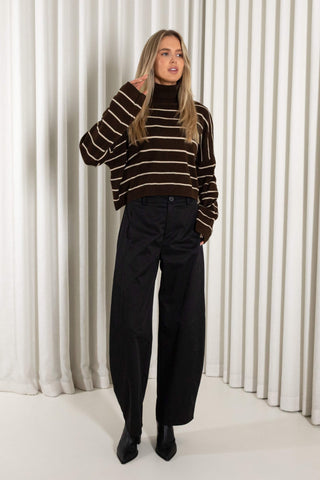 A woman wearing a cropped brown knit turtleneck sweater with horizontal cream stripes and black wide-leg pants, standing against a white pleated curtain backdrop.