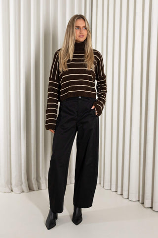 A woman wearing a cropped brown knit turtleneck sweater with horizontal cream stripes and black wide-leg pants, standing against a white pleated curtain backdrop.