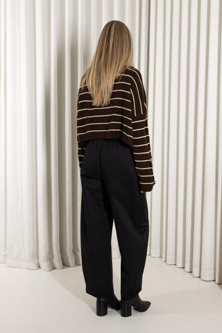 A woman wearing a cropped brown knit turtleneck sweater with horizontal cream stripes and black wide-leg pants, standing against a white pleated curtain backdrop.