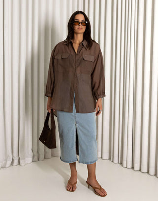 Woman wearing a brown shirt and blue jeans standing against a white curtain background Brown sheer long sleeve collared button-down shirt