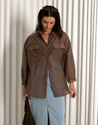 Woman wearing a brown shirt and blue jeans standing against a white curtain background Brown sheer long sleeve collared button-down shirt