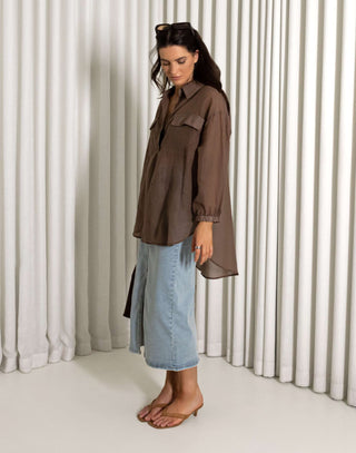 Woman wearing a brown shirt and blue jeans standing against a white curtain background Brown sheer long sleeve collared button-down shirt