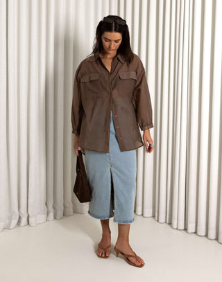 Woman wearing a brown shirt and blue jeans standing against a white curtain background Brown sheer long sleeve collared button-down shirt