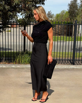 A woman stands outdoors looking at her phone and wearing a black off shoulder top and midi skirt, holding a black clutch.