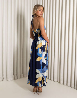 Woman wearing a blue floral dress against a white curtain background Blue midi dress with oversized floral print and tie halter neckline

