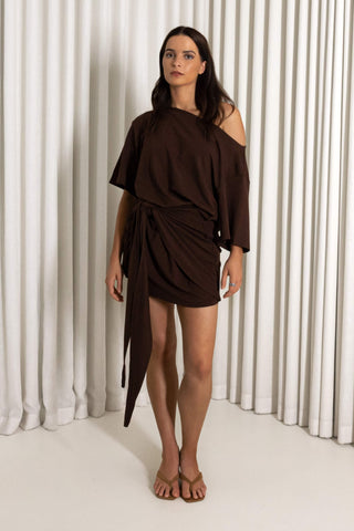 A woman modeling a dark brown off-the-shoulder wrap mini dress with a draped waist tie, standing against a white pleated curtain backdrop.