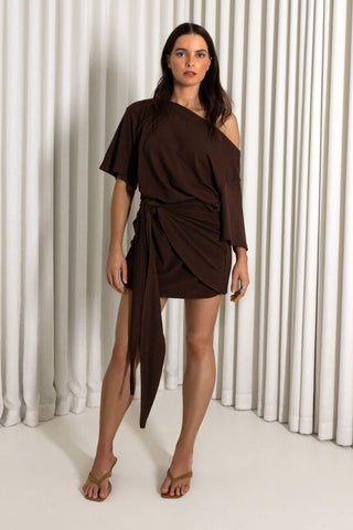 A woman modeling a dark brown, one-shoulder wrap mini dress with a side slit and tie belt, standing against a white curtain backdrop.