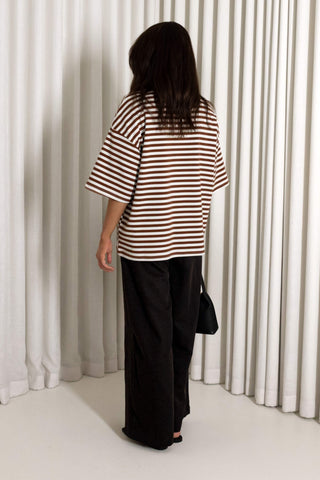 A woman viewed from behind wearing an oversized brown and white striped short-sleeve top, black wide-leg pants, and holding a black handbag in front of white pleated curtains.