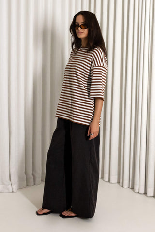A woman modeling an oversized brown and white horizontally striped short-sleeve top with black wide-leg pants and black sandals, standing in front of white pleated curtains.