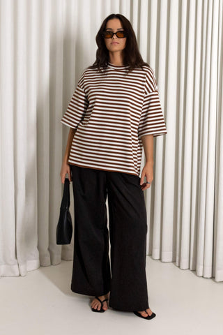 A woman modeling an oversized brown and white horizontally striped short-sleeve t-shirt paired with black wide-leg pants and black sandals, standing in front of a white pleated curtain backdrop.