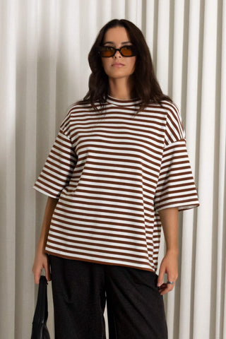 A woman wearing an oversized brown and cream striped t-shirt with elbow-length sleeves, paired with black pants, standing in front of a white pleated curtain backdrop.