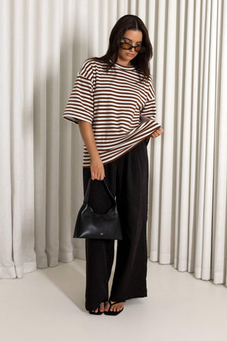 A woman modeling an oversized brown and white striped t-shirt, black wide-leg pants, and a black leather handbag, standing in front of white pleated curtains.