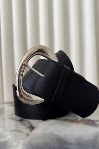 A black leather belt with a large, polished silver-tone oval buckle, coiled on a marble surface with a soft-focus background of vertical blinds.