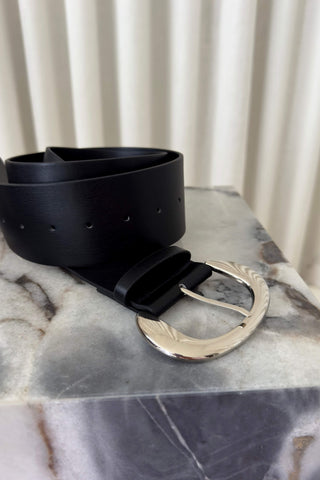 A black leather belt with a large, polished silver-tone oval buckle resting on a gray and white marble block, with a cream-colored pleated curtain in the background.