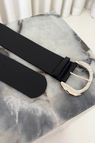 A black leather belt with a polished silver-tone metal buckle, displayed diagonally on a gray and white marble surface.