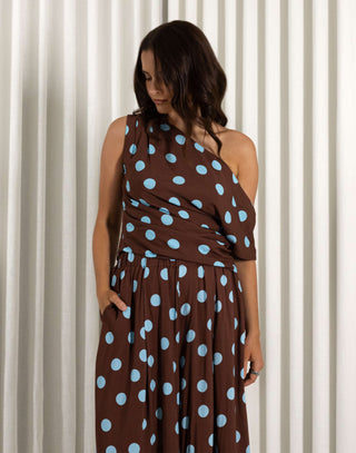 Woman wearing a brown top and matching pants with blue polka dots against a white curtain background. Brown off shoulder top with gathered detailing and relaxed fit