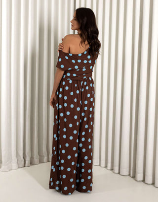 Woman wearing a brown top and matching pants with blue polka dots against a white curtain background. Brown off shoulder top with gathered detailing and relaxed fit