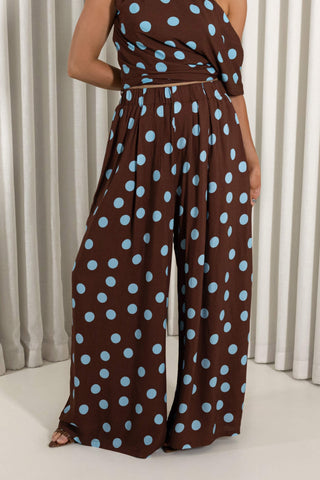 A woman wearing a brown one-shoulder jumpsuit with light blue polka dots, paired with a matching waist tie, set against a neutral curtain background.