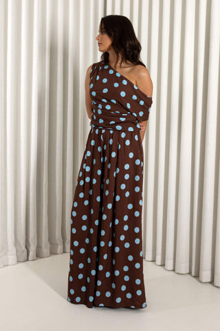 A woman modeling a brown one-shoulder maxi dress with light blue polka dots, standing in front of a white pleated curtain backdrop.