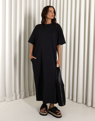 Woman wearing a black dress standing against a white curtain background Black relaxed fit midi dress with side leg split