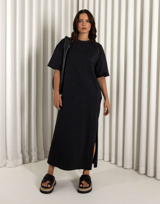 Woman wearing a black dress standing against a white curtain background Black relaxed fit midi dress with side leg split