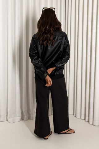 A woman stands with her back to the camera wearing a black faux-leather jacket, black wide-leg pants, and black sandals in front of white pleated curtains.