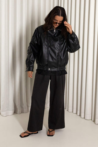 A model wearing a black faux-leather jacket, black wide-leg pants, and black flip-flops, standing in front of a white pleated curtain backdrop.
