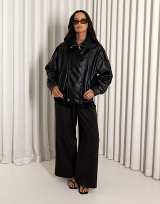 Person wearing a black oversized faux leather bomber and pants standing against a white curtain.