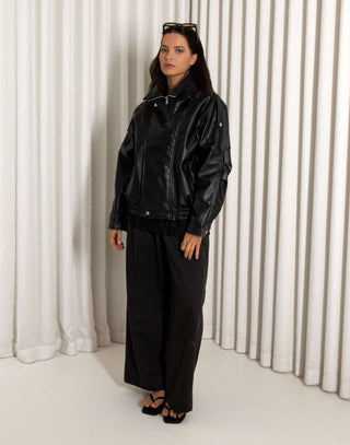 Person wearing a black oversized faux leather bomber and pants standing against a white curtain.