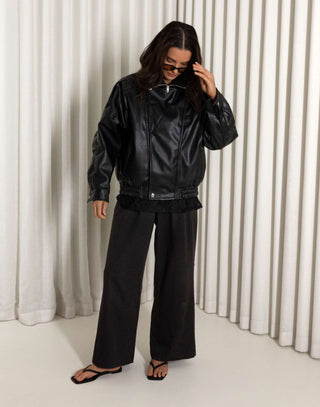 Person wearing a black oversized faux leather bomber and pants standing against a white curtain.