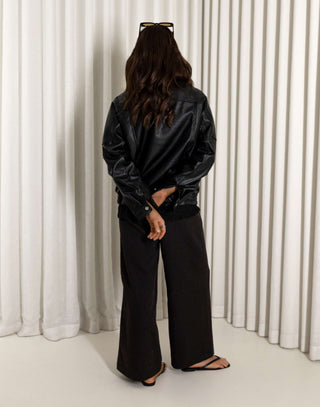 Person wearing a black oversized faux leather bomber and pants standing against a white curtain.