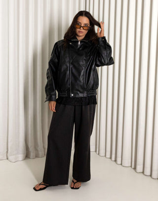Person wearing a black oversized faux leather bomber and pants standing against a white curtain.